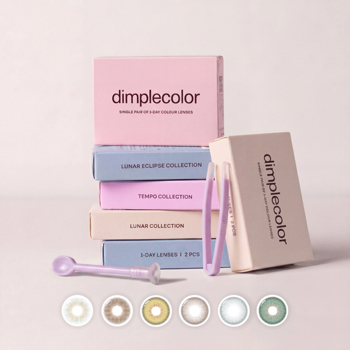 A set of Dimplecolor contact lens boxes and accessories arranged on a light beige background. The stack includes pink, blue, lavender, and cream boxes labeled with Collection names and 1-Day lenses, with a small applicator tool and tweezers.